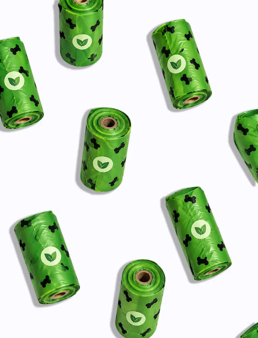 Several rolls of green plastic bag liners arranged on a white background. Each roll has a white oval logo with a green leaf and small black dog-bone motifs printed across it.