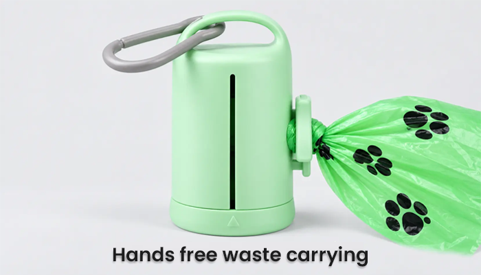 A mint green portable pet waste bag dispenser with a gray carabiner clip, shown dispensing a green plastic bag printed with black paw prints. The text 'Hands free waste carrying' is displayed at the bottom.