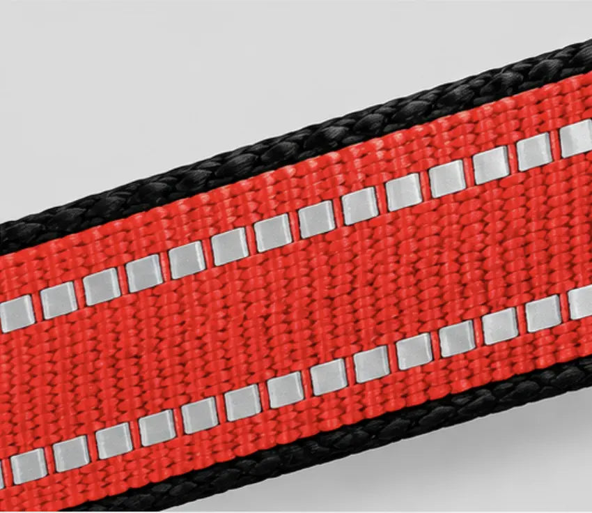 Close-up of a red woven reflective strap with black braided edges and silver reflective squares running parallel to the center, shown on a light gray background.