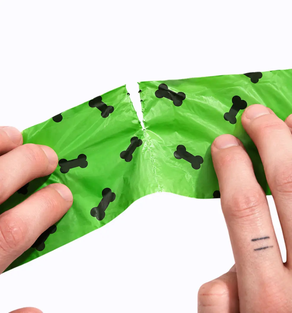 Hands tearing a green LuxryPaw dog waste bag with a black bone pattern along the perforated edge