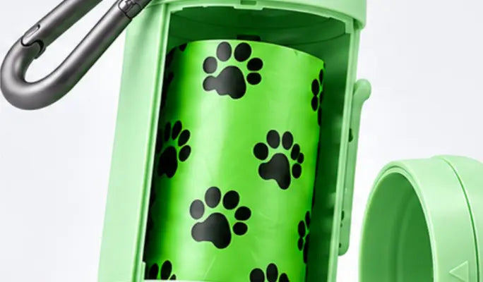 A close-up of a light green pet poop bag holder with a black carabiner clip and a roll of poop bags printed with black paw prints, shown against a white background.